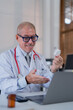 © MINAE - Senior male doctor in a white coat and glasses engaging in a virtual consultation, holding a medicine bottle, demonstrating telemedicine practices in a modern office setting