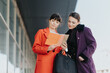 © qunica.com - Two women in stylish coats engage in a business discussion while examining notes, showcasing teamwork and collaboration in a casual outdoor setting.