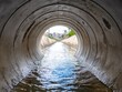 © Netsai - View from inside a large concrete pipe, with water flowing towards the opening, revealing urban landscape in the distance.