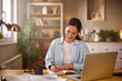 © Stockphotodirectors - A woman sits at her desk in a home office, writing in a notebook with a laptop in front of her. She enjoys a cup of coffee, surrounded by plants and cozy decor.