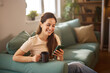 © Stockphotodirectors - A young woman sits comfortably on a couch, sipping coffee from a mug while engaged with her smartphone. The living room is filled with natural light and plants.