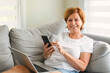 © Louis-Paul Photo - senior woman relaxing at home on the sofa reading her text messages on her mobile phone with a quiet smile