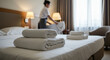© EPICreation - Portrait of hotel room with fresh white towels on bed and housekeeper making the other bed in the background - hospitality and comfort concept