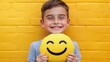 © Oleksandr - A cheerful young person holds a bright yellow emoji pillow with a smiling face, standing against a vibrant yellow wooden background with a joyful, playful expression.