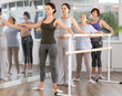 © JackF - Concentrated graceful female ballet instructor leading rehearsal at barre with group of amateur women of various ages in choreography studio, practicing basic second position