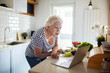 © Marko Geber - Senior woman looking up recipes on laptop while preparing fresh vegetables in kitchen