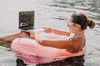 © svetograph - A woman is sitting in a pink inflatable raft on a lake, using a laptop. Concept of relaxation and leisure, as the woman is enjoying her time outdoors while working on her laptop.