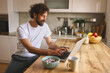 © Stockphotodirectors - A man is sitting at a wooden kitchen table, typing on his laptop while having breakfast. He has a bowl of cereal and a cup of coffee beside him, showing a relaxed morning ambiance.
