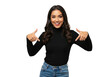 © Over View - A smiling young woman with long dark hair points down with both thumbs, wearing a black turtleneck and jeans, isolated on a transparent background