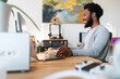 © Studio Marmellata - An African American man smiles broadly while working on his laptop and computer monitor at a bright, modern office desk. He is focused and engaged in his professional tasks.