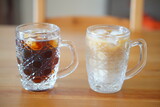 透明なガラス製のジョッキに入った二種類の冷たい飲み物が、温かみのある木製のテーブルの上に並べられた、涼しげな一枚。左のジョッキにはアイスコーヒー、右にはアイスカフェオレがそれぞれ氷と共に注がれており、夏の日差しの中で喉の渇きを癒すような、爽やかなひとときを演出している。