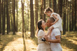 © aleonovs - A joyous child on father's shoulders, her mother is nearby looking up, all smiling in a sun-drenched pine forest