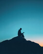 © Kowit - Silhouette of a person sitting on a rock, reading against a clear twilight sky with a gradient from blue to orange.