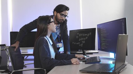 Diverse IT professionals collaborating in a command center. A male programmer explains data on a tablet to a female developer working on code at her computer.
