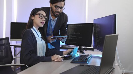 Diverse IT professionals collaborating in a command center. A male programmer explains data on a tablet to a female developer working on code at her computer.