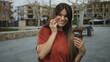 © Krakenimages.com - Young hispanic woman talking on smartphone while holding coffee cup outdoors on city street with blurred urban background.