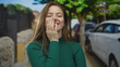 © Krakenimages.com - Young smiling woman in a green sweater covers half her face with her hand on a sunlit urban street; shyness.