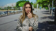 © Krakenimages.com - Woman wearing striped shirt looking concerned standing on street outdoors in sunny urban area with green trees and buildings in the background.