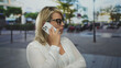 © Krakenimages.com - Young blonde woman wearing glasses and white blouse holds smartphone to ear on busy city street in daylight; confidence.