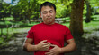 © Krakenimages.com - Man in red shirt clutches stomach painfully in a sunny park surrounded by lush greenery, highlighting discomfort outdoors.