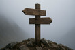 © Koushik - Old wooden direction signpost with two arrows on foggy mountain trail creating concept of decision crossroads and travel choice during moody misty adventure hike