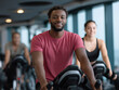 © alsu0112 - Young African American man smiling while exercising on stationary bike in modern gym, with fitness enthusiasts in background, promoting healthy lifestyle and community engagement