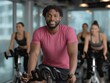 © alsu0112 - African American man in pink shirt is smiling while cycling on stationary bike in modern gym with women exercising in background, showcasing fitness and motivation