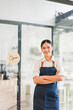 © kenchiro168 - Young female barista standing confidently with arms crossed inside cafe near open sign, smiling warmly and ready to serve customers in casual work apron