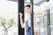 © kenchiro168 - Young woman smiling while opening glass door of store with open sign, welcoming customers with friendly expression