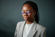 © Seventyfour - Portrait of young adult Black woman wearing eyeglasses and jewelry looking at camera with confident expression, standing against plain background in professional attire