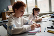 © Seventyfour - Caucasian boy and Caucasian girl sitting at desk writing in notebooks during classroom lesson, focused on learning, school supplies scattered on table, sunlight streaming through window blinds