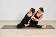 © TRAVELARIUM - Female yoga instructor guides adult woman in seated stretch on yoga mat. Minimalist studios neutral tones highlight focus and calm. Both wear black outfits, emphasizing form and balance
