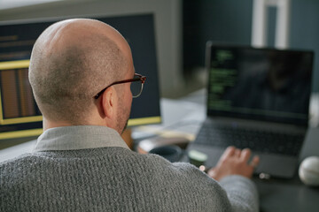 middle aged caucasian man working on laptop with code visible on screen, sitting at desk in modern o