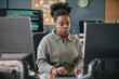 © Seventyfour - Black young adult woman working at computer desk, focusing on screen while typing on keyboard, surrounded by multiple monitors in modern office setting with code visible in background