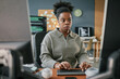 © Seventyfour - Young adult Black woman working at computer desk, focusing on typing with hands on keyboard, sitting in modern office environment with multiple monitors and programming code visible