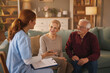 © Stockphotodirectors - A home health aide, wearing scrubs and holding a clipboard, speaks to a senior couple sitting on a couch in their living room, discussing details of their care and needs.