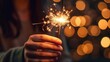 © AlexTroi - Close-up of a hand holding a glowing sparkler with golden bokeh lights in the background, symbolizing celebration, warmth, holiday cheer, and festive ambiance.