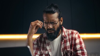 Indian male programmer suffering eyestrain while coding on laptop from home