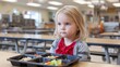 © Oleksandr - A young child with blonde hair sits at a table with a cafeteria tray of food, looking thoughtful in a school or institutional dining area.