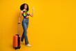 © deagreez - Stylish young woman with curly black hair waving while rolling an orange suitcase against vibrant yellow background
