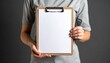 © photostockatinat - Person holding a clipboard with a blank sheet of paper. The composition suggests a professional or administrative context. The person is wearing a t-shirt.