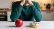© kelum - Man contemplating healthy eating choices with apple versus donut on a kitchen table, making a difficult lifestyle decision