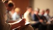 © AspctStyle - Focused individual taking notes with a pen on a clipboard during a seminar or business conference, with a blurred row of seated attendees in the background under warm, professional lighting