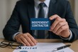 © Generative AI - Businessman in formal suit holding independent contractor card with work documents on table.