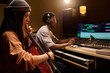 © pressmaster - Latin girl playing cello while Black young adult man wearing headphones working at mixing console in music studio, both focused on creative music production process together