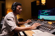 © pressmaster - Young adult Black man wearing headphones operating audio mixing console and computer monitors in professional recording studio, adjusting sound levels and working with music production equipment