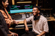 © pressmaster - Young Black man sitting in music studio wearing headphones around neck, smiling and interacting with young Hispanic woman while surrounded by audio mixing equipment and computer monitors