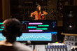 © pressmaster - Young adult Latin woman playing cello in professional recording studio, sitting behind microphone with music producer monitoring audio levels on mixing console in foreground