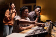 © pressmaster - Young Black man operating audio mixing console in recording studio, smiling while young Black man and young Hispanic woman standing nearby, woman holding smartphone and capturing moment