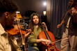 © pressmaster - Latin girl playing cello surrounded by Black young men playing violins in music studio, focused expressions, musical collaboration, instruments visible in background